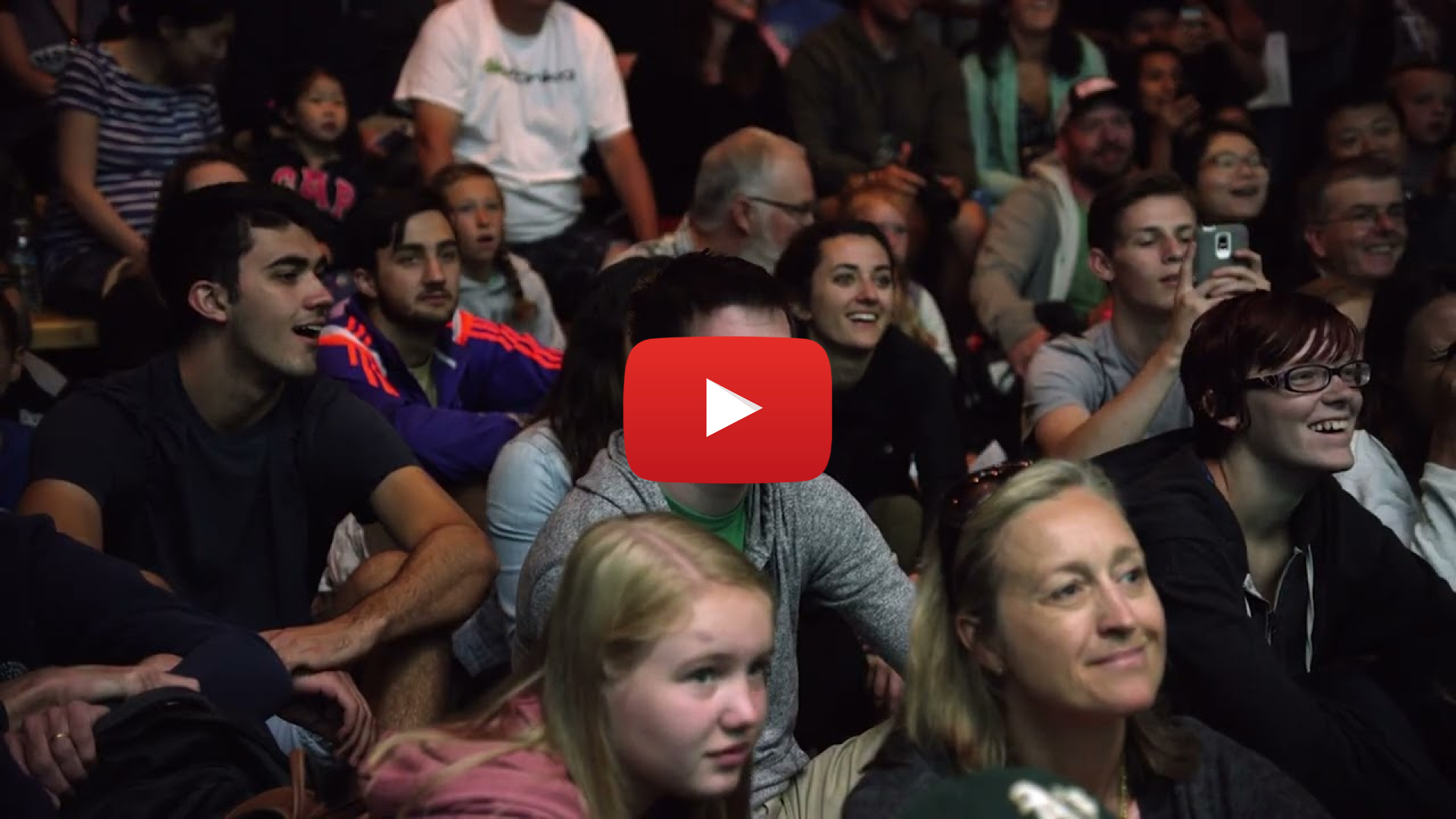 A closeup of a smiling audience. Overlaid is a video play button.