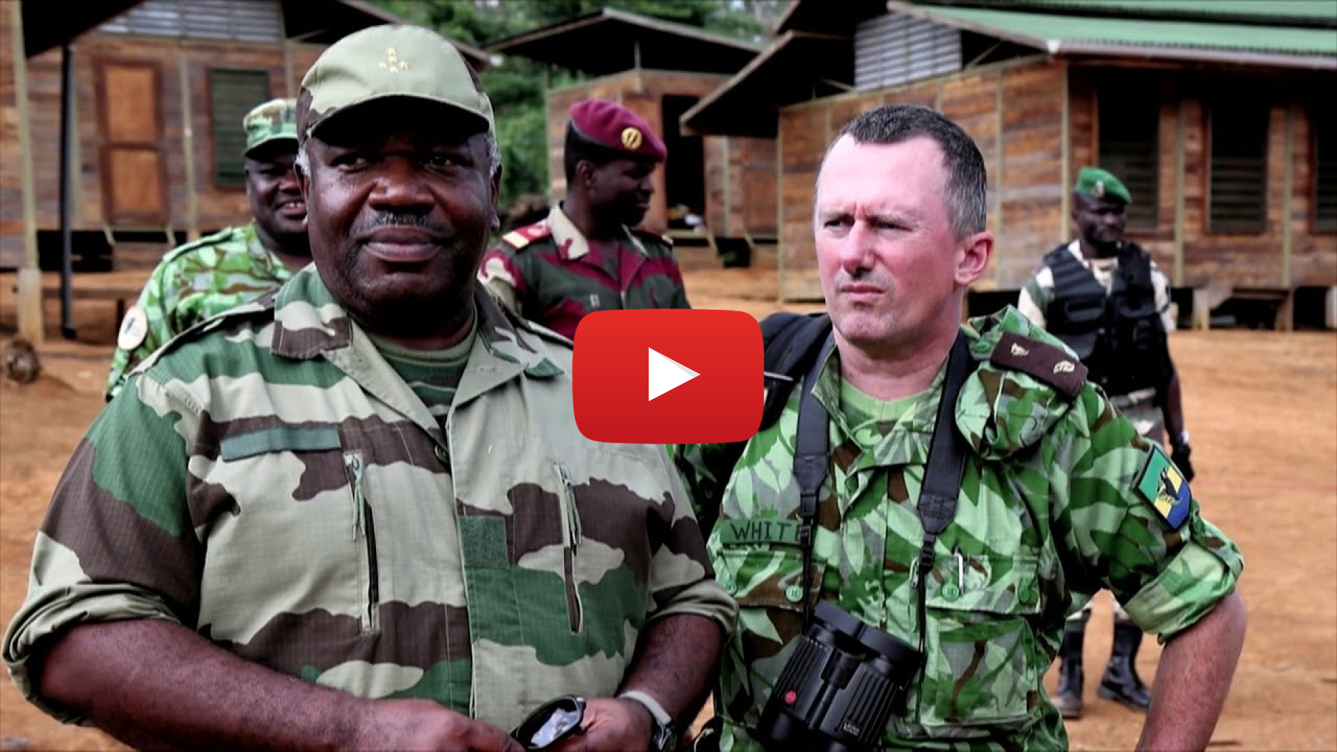 Two men in camouflage fatigues in front of a small village. Overlaid is a video play button.