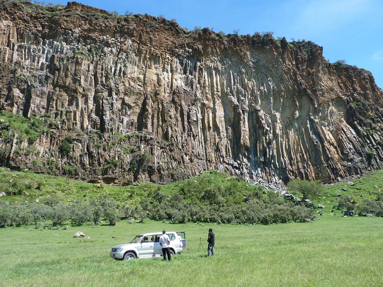 Several scientists standing next to a vehicle and looking up at a steep rocky cliff.