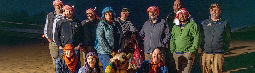 A group of scientists in desert wear at night posing and smiling next to a camel