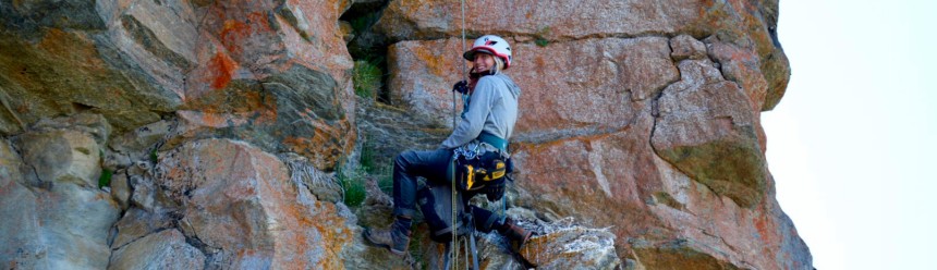 Biologist Kara Beer in climbing gear on the side of a rocky cliff smiling at the camera.