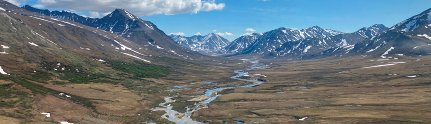 An aerial view of an Arctic landscape with snow-covered mountains and a river coursing through a green valley.