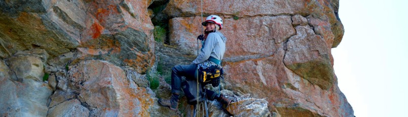 Biologist Kara Beer in climbing gear on the side of a rocky cliff smiling at the camera.