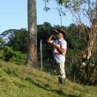 A man standing with troptical trees behind him holding a clipboard and looking at something through binoculars