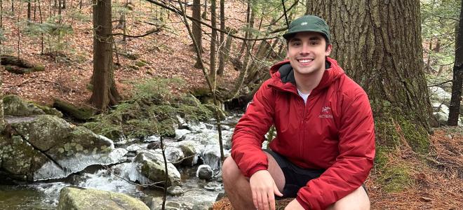 Luke Carmosino squatted in a forest, smiling for a photo