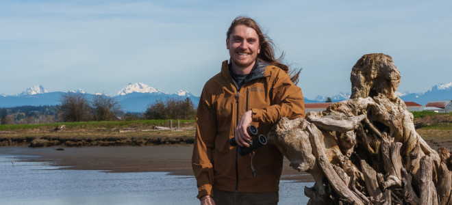 Austin Breuer posing in front of a dead tree stump with water, an open landscape, and distant snow-covered mountains behind him.
