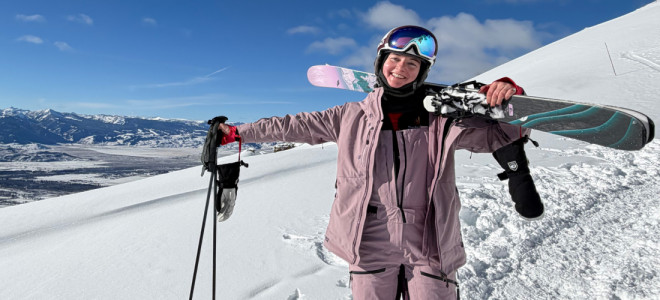 Katy Axel in skiing gear and carrying skis posing and smiling on a snow-covered hill with distant mountains behind her.