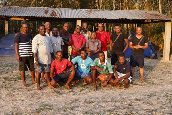 15 men, all part of our field team, stand together in front of roofed structure with tents underneath