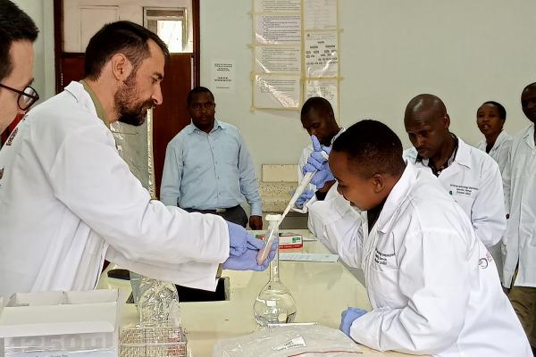 People in lab coats gathered around as one of them uses a syringe to put a liquid into a vile filled with a pink liquid