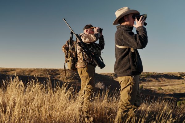 Two hunters standing in a grassy field looking through binoculars