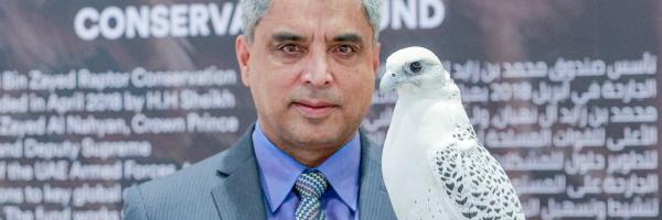 Munir Virani, ex-Vice President of The Peregrine Fund, poses in front of Mohamed Bin Zayed Raptor Conservation Fund banner, while holding a Gyrfalcon