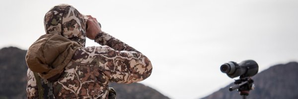 A scientist seen from behind standing next to a spotting scope and using binoculars to look at distant hills