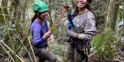 Two smiling biologists wearing climbing gear standing at the base of a tree