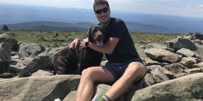 A seated man holds a dog close in a field of boulders during a hike