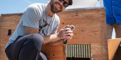 A man on one knee, poses for photo with a baby bird.