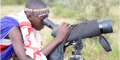 A young person in bright, traditional clothing of the Maasai tribe of Kenya looking through a spotting scope