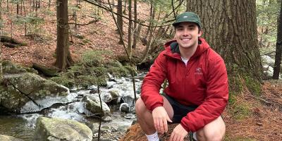 Luke Carmosino squatted in a forest, smiling for a photo