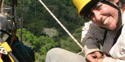 A smiling biologist in climbing gear high in a tree with treetops seen far below her in the background