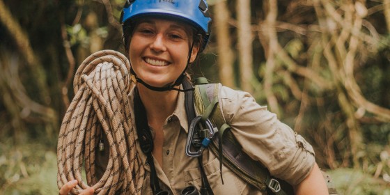 Hana in the field wearing climbing gear and carrying a coiled rope