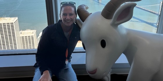Stacie Johnson posing and smiling next to a statue of a goat with a scenic view of city buildings and a suspension bridge over water in the background.