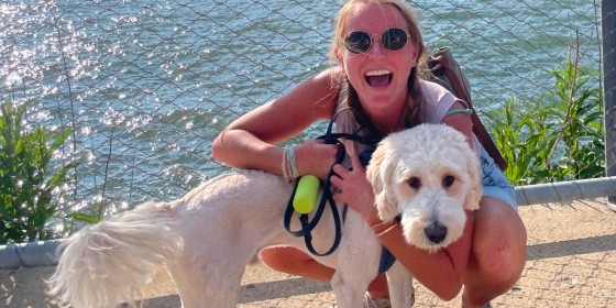 Tali Brennan posing and laughing while kneeling down with her arms around a fluffy white dog.
