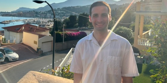 William Gingold posing and smiling with a scenic view of rolling hills, a harbor, and a quaint village behind him.
