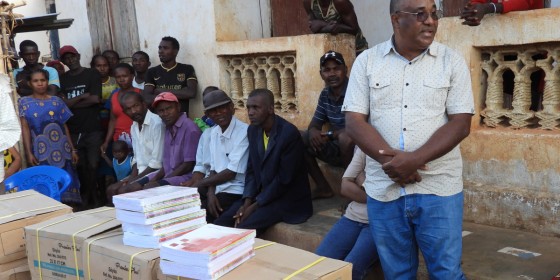 Madagascar Program director Lily-Arison Rene de Roland speaking to a community gathering and standing behind pallets of school supplies.