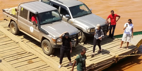 Two vehicles and a small group of people standing on a wooden ferry boat in a muddy river.