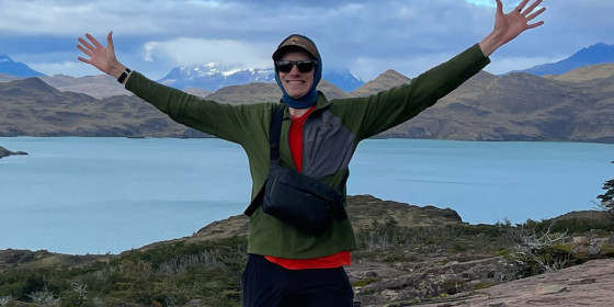 Burr Tweedy standing with his arms raised in front of a lake and distant snow-capped mountains.