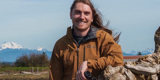 Austin Breuer posing in next to a tree stump with water, an open landscape, and snow-capped mountains visible behind him.